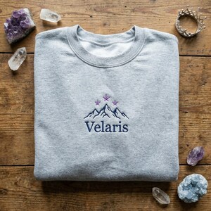 May include: A light gray sweatshirt with the embroidered word "Velaris" below a mountain and star design in navy blue and purple. The sweatshirt is folded neatly on a wooden surface.