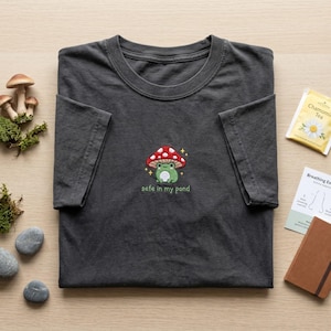 Könnte beinhalten: Dunkelgraues T-Shirt mit einer Stickerei einer grünen Froschfigur unter einem rot-weiß gepunkteten Pilz und dem Text "safe in my pond". Das T-Shirt ist gefaltet. Daneben liegen ein Teebeutel und ein Notizbuch.