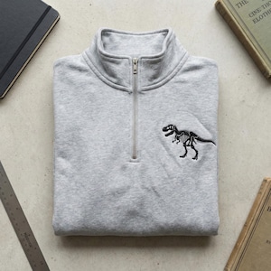 May include: A light gray sweatshirt with a quarter-zip collar and a black embroidered dinosaur skeleton design on the chest. The sweatshirt is folded neatly, showcasing its details.
