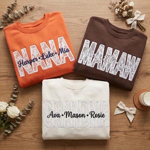 May include: Three crewneck sweatshirts in orange, brown, and cream. Each features large block letters filled with white lace fabric. The orange sweatshirt says NANA with names Harper, Luke, and Mia. The brown says MAMA with Avery, Ellie, and Cooper. The cream says GRANDMA with Ava, Mason, and Rosie.