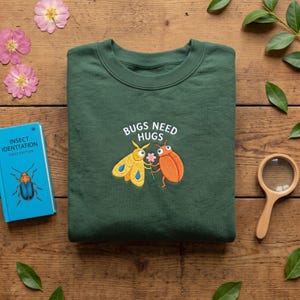 May include: A dark green crewneck sweatshirt featuring an embroidered graphic of a yellow moth and an orange beetle holding a small pink flower. Above the insects, the white text reads "BUGS NEED HUGS" in a simple, clean, sans-serif font.