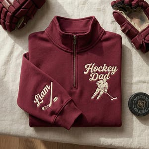 May include: A burgundy hockey-themed sweatshirt with a quarter-zip neckline. The text "Hockey Dad" and a hockey player graphic are embroidered on the front. The sleeve features the name "Liam" and a hockey stick and puck.