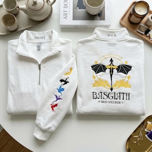 May include: White sweatshirt with a quarter-zip collar. The front of the sweatshirt has the word "FRONT" printed on the collar. The left sleeve features embroidered colourful dragons. The back has a dragon emblem and the words "BASGIATH + WAR COLLEGE+".