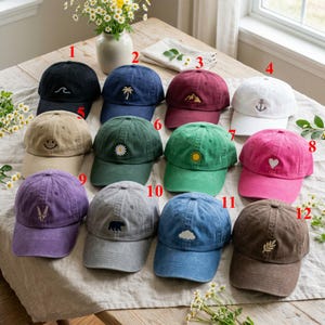 Könnte beinhalten: Eine Auswahl von zwölf Baseballkappen in verschiedenen Farben, jede mit einem einzigartigen gestickten Design. Farben sind Schwarz, Marineblau, Burgunderrot, Weiß, Beige, Grün, Rosa, Lila, Grau und Braun. Designs umfassen Welle, Palme, Berg, Anker, Smiley, Gänseblümchen, Sonne, Herz, Weizen, Bär und Wolke.