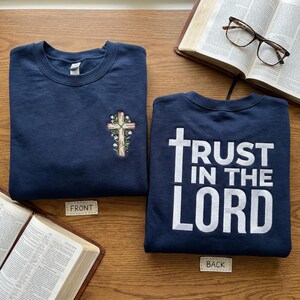 May include: Two navy blue sweatshirts are shown. One has an embroidered cross with floral details. The other displays the white text "TRUST IN THE LORD". Open books and spectacles are in the background.