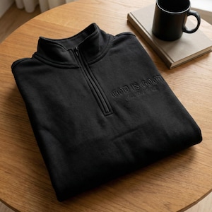 Könnte beinhalten: Ein schwarzes Sweatshirt mit Viertelreißverschluss und dem gestickten Text "GOD IS GOOD ALL THE TIME". Das Sweatshirt liegt gefaltet auf einem Holztisch, im Hintergrund eine schwarze Tasse und ein Buch.
