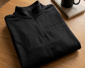 May include: A black quarter-zip sweatshirt with the embroidered text "GOD IS GOOD ALL THE TIME." The sweatshirt is folded on a wooden table, with a black mug and a book in the background.