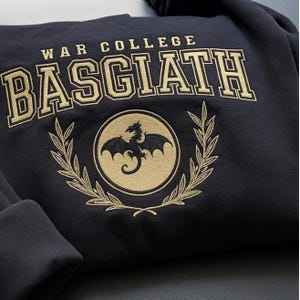 May include: Navy blue sweatshirt with gold embroidered text and design. The text reads "WAR COLLEGE BASGIATH" above a circular emblem featuring a dragon and laurel wreath. A cozy, casual garment.