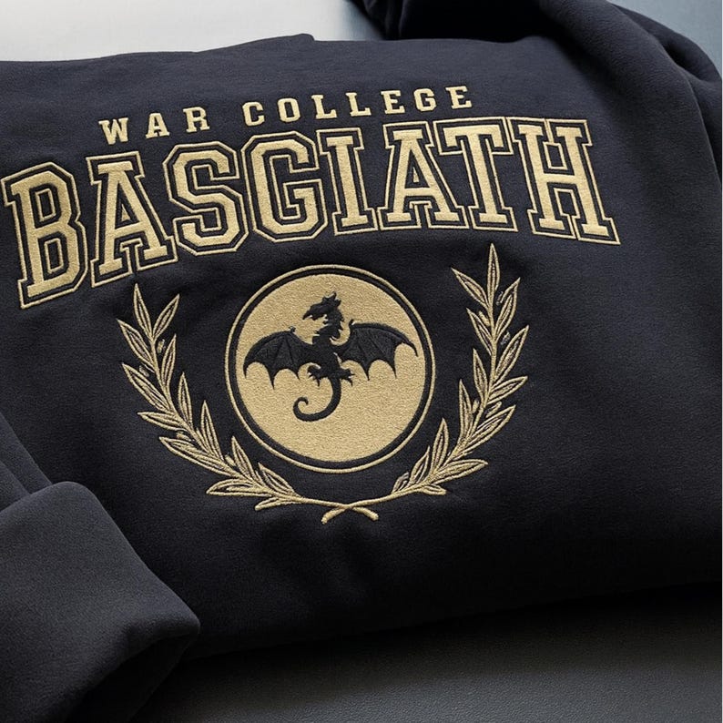 May include: Navy blue sweatshirt with gold embroidered text and design. The text reads "WAR COLLEGE BASGIATH" above a circular emblem featuring a dragon and laurel wreath. A cozy, casual garment.