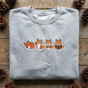 Könnte beinhalten: Ein meliertes graues Sweatshirt mit einem gestickten Design von vier Cartoon-Füchsen. Die Füchse werden in verschiedenen Posen dargestellt: schlafend, winkend, eine Tasse haltend und ein Buch lesend. Die Füchse sind orange und weiß.