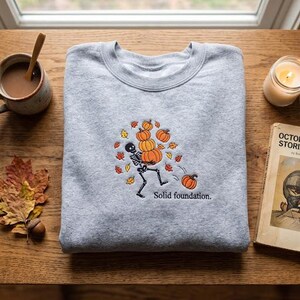 May include: A light gray crewneck sweatshirt featuring an embroidered design of a skeleton juggling orange pumpkins surrounded by colorful autumn leaves. Below the skeleton, the text reads Solid foundation. in black, simple, lowercase lettering.
