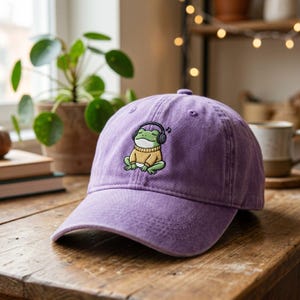 Può includere: Un cappellino da baseball lilla con un ricamo di un ranocchio dei cartoni animati con cuffie e un maglione giallo. Il ranocchio è seduto con le gambe incrociate. Il cappellino è realizzato in un tessuto morbido e strutturato.