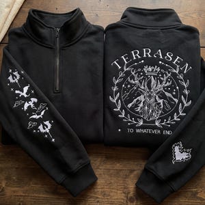 May include: A black quarter-zip sweatshirt featuring white embroidery. The sleeve displays flying dragons and stars. The back features a geometric stag head, a crown, and the text TERRASEN TO WHATEVER END surrounded by a laurel wreath.