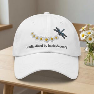 May include: White baseball cap featuring a daisy chain and a dragonfly design. The text "Radicalized by basic decency" is printed below the daisy chain. The cap is made of a soft material.