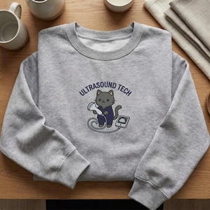 May include: A grey sweatshirt with a cartoon cat in a navy blue uniform holding an ultrasound probe. The text "ULTRASOUND TECH" is above the cat. The cat is connected to a small monitor via a cable.