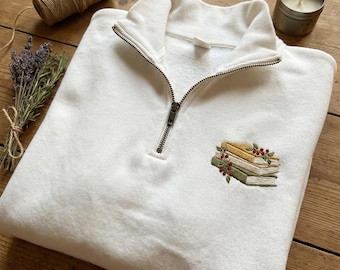 May include: A white quarter-zip sweatshirt with an embroidered stack of books in shades of green, yellow, and brown, accented with red berries. The sweatshirt has a ribbed collar and cuffs, and a metal zipper.