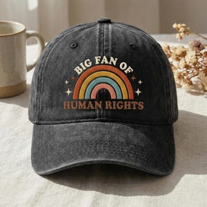 May include: A dark grey baseball cap with the text "BIG FAN OF HUMAN RIGHTS" above a rainbow graphic. The cap has a curved peak and is made of a worn fabric. The rainbow design includes orange, yellow, blue, and pink colours.