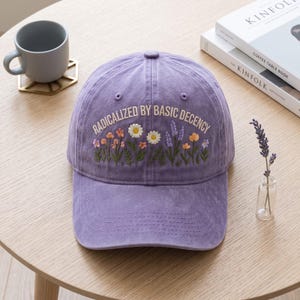 May include: A purple baseball cap featuring embroidered wildflowers and the text RADICALIZED BY BASIC DECENCY in white lettering. The cap has a curved brim, a button on top, and stitched eyelets on the crown, showcasing a soft, washed fabric texture.