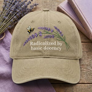 Op de afbeelding: Een kaki baseballcap geborduurd met een bij en lavendeltakjes. De tekst "Radicalized by basic decency" is geborduurd onder het bloemmotief. De hoed ligt op een houten oppervlak.