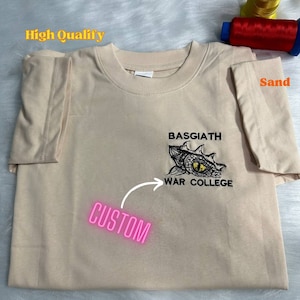May include: A sand-coloured t-shirt with a black embroidered design of a dragon's eye and the text "BASSGIATH WAR COLLEGE". The text "CUSTOM" is written in pink on the shirt.