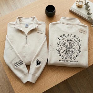 May include: Two cream-coloured quarter-zip sweatshirts. One shows the front with a zip and the text "You do not fear. You do not falter. You do not yield." on the sleeve. The other displays the back with the text "TERRASEN HOME OF THE STAG" and a stag illustration.