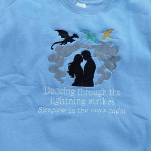May include: Light blue sweatshirt with embroidered design. Features a silhouette of a couple, dragons, and text that reads "Dancing through the lightning strikes" and "Sleepless in the onyx night."