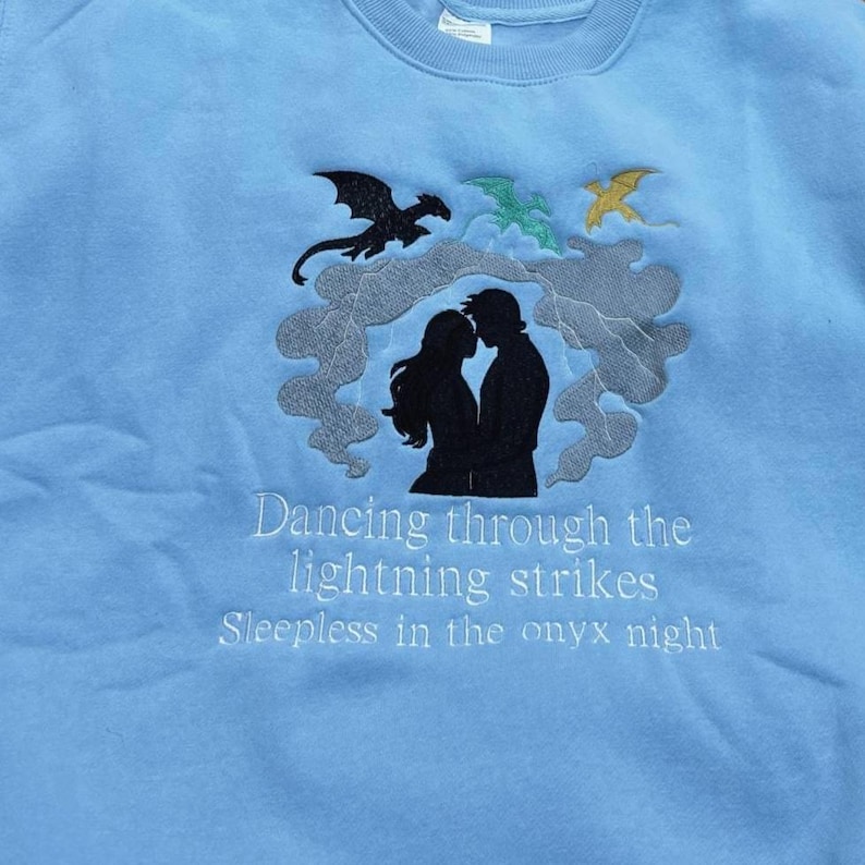 May include: Light blue sweatshirt with embroidered design. Features a silhouette of a couple, dragons, and text that reads "Dancing through the lightning strikes" and "Sleepless in the onyx night."