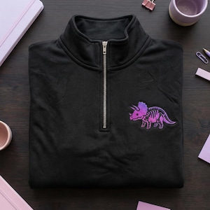 May include: Black quarter-zip sweatshirt with a purple and pink triceratops patch on the chest. The dinosaur patch features a gradient design. The sweatshirt has a high collar and a silver zipper.