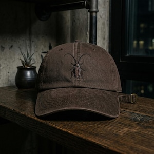 May include: A brown baseball cap with a stitched cockroach design. The cap has a curved brim and an adjustable strap. The cockroach is embroidered in a detailed, realistic style.