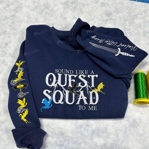 May include: A navy blue long-sleeved sweatshirt with the text "SOUND LIKE A QUEST SQUAD TO ME" embroidered in white. The sweatshirt has yellow and blue dragon designs embroidered on the sleeves.