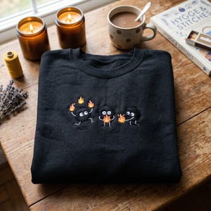May include: Black sweatshirt with embroidered cartoon characters holding flames. The sweatshirt is folded on a wooden surface, with lit candles, a cup of coffee, and a book in the background.