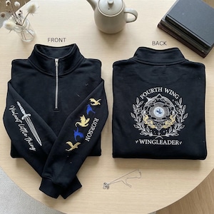 May include: Black quarter-zip sweatshirt with embroidered details. The front features a sword and text "Violent Little Thing" on the sleeve. The other sleeve has embroidered dragons and the word "Rioroson". The back displays "Fourth Wing" and "Wingleader".