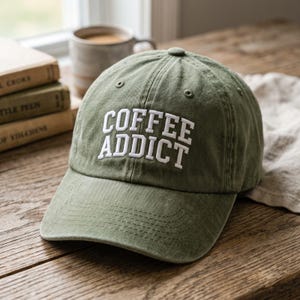 May include: An olive green baseball cap featuring the white embroidered text COFFEE ADDICT on the front panel. The cap has a structured crown, a curved brim with tonal stitching, and small ventilation eyelets near the top button.