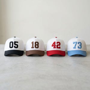 Könnte beinhalten: Vier weiße Baseballkappen mit unterschiedlich farbigen Schirmen und Zahlen. Die Kappen haben schwarze, braune, rote und hellblaue Schirme. Jede Kappe hat eine andere Zahl in einer Kontrastfarbe: 05, 18, 42 und 73.