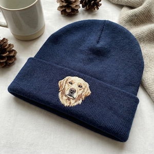 May include: A navy blue beanie featuring an embroidered golden retriever dog. The hat is made of a soft knit material and has a folded brim. The dog's face is detailed with brown and tan thread.