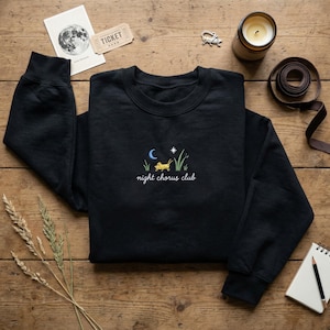 May include: Black crewneck sweatshirt with embroidered design. The design features a crescent moon, star, grass, and a grasshopper, with the words "night chorus club" in cursive. The sweatshirt is displayed on a wooden surface with other items.