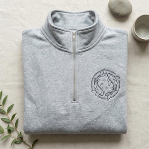 Könnte beinhalten: Hellgraues Sweatshirt mit Reißverschluss und hohem Kragen. Das Sweatshirt hat einen silbernen Reißverschluss und ein gesticktes rundes Design mit zwei Fischen und Sternen. Ein kleiner Zweig mit Grün, ein Stein und eine kleine Keramikschale befinden sich im Hintergrund.