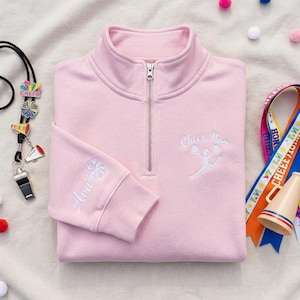 Könnte beinhalten: Hellrosa Sweatshirt mit Viertelreißverschluss, bestickt mit den Worten "Cheer Mom" und einer Cheerleader-Grafik in Weiß. Der Ärmel hat "Ava" und einen Pompon, ebenfalls weiß. Cheerleading-Zubehör ist im Hintergrund zu sehen.