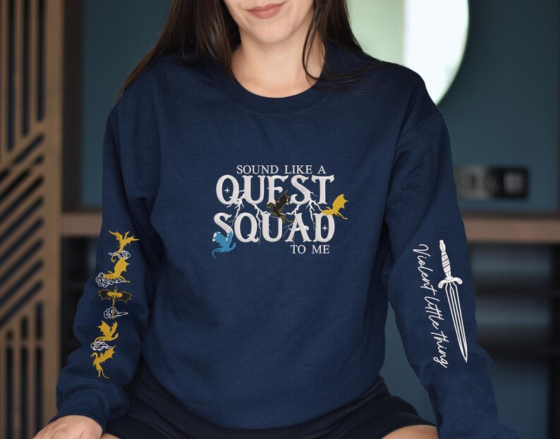 May include: A navy blue long-sleeved shirt with a white graphic that says "Sound like a Quest Squad to me". The graphic includes a lightning bolt and stylized dragons. The left sleeve has a repeating pattern of stylized dragons. The right sleeve has a white graphic of a sword with the text "Violent Little Thing".
