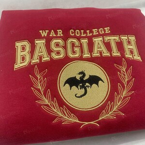 May include: A red sweatshirt with gold embroidered text and a circular emblem. The text reads "WAR COLLEGE BASGIATH". The emblem features a black dragon within a gold circle, surrounded by a gold laurel wreath.