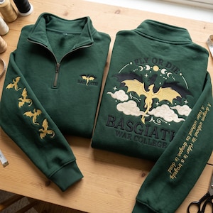 May include: Two dark green quarter-zip sweatshirts featuring gold and black dragon embroidery. The front displays a small dragon and "BASGIATH". The back features a large dragon, clouds, moon phases, and "BASGIATH WAR COLLEGE".