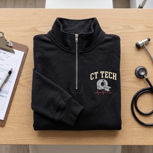 May include: A black quarter-zip sweatshirt featuring an embroidered design on the chest. The embroidery includes the text "CT TECH" in beige letters above a gray CT scanner icon and a red EKG heart rhythm line. The sweatshirt has a silver zipper.