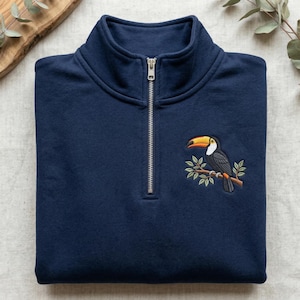 Peut inclure: Sweat-shirt bleu marine à fermeture éclair quart de zip avec une broderie d'un toucan sur une branche. Le toucan a un bec jaune et orange, un corps noir et des détails blancs. Col montant et fermeture éclair argentée.