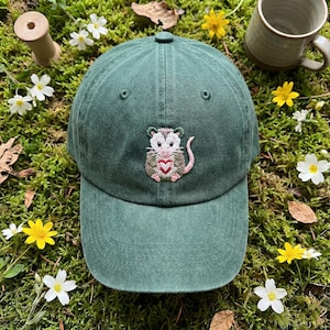 K&ouml;nnte beinhalten: Eine waldgr&uuml;ne Baseballkappe mit einer gestickten Grafik eines kleinen Opossums, das ein rot-wei&szlig;es Herz h&auml;lt. Die Kappe verf&uuml;gt &uuml;ber eine strukturierte Krone, einen gebogenen Schirm und einen Knopf oben, mit detaillierten Stickereien.
