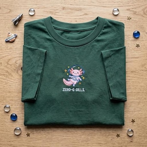 May include: A dark green t-shirt featuring an embroidered graphic of a pink axolotl wearing a space helmet and suit, floating among small yellow stars. Below the axolotl, the text reads ZERO-G GILLS in white, capitalized, sans-serif letters.