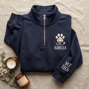 May include: Navy blue quarter-zip sweatshirt with a white paw print design and stethoscope, personalized with the name "ISABELLA." The sleeve features the text "VET TECH" in white. The sweatshirt is folded and displayed on a neutral surface.
