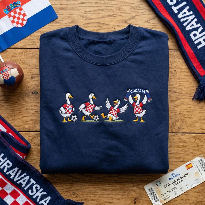 May include: A navy blue crewneck sweatshirt featuring an embroidered graphic of four white geese wearing red and white chequered jerseys. The geese are playing football, and one holds a blue and red scarf with the text CROATIA printed in white.