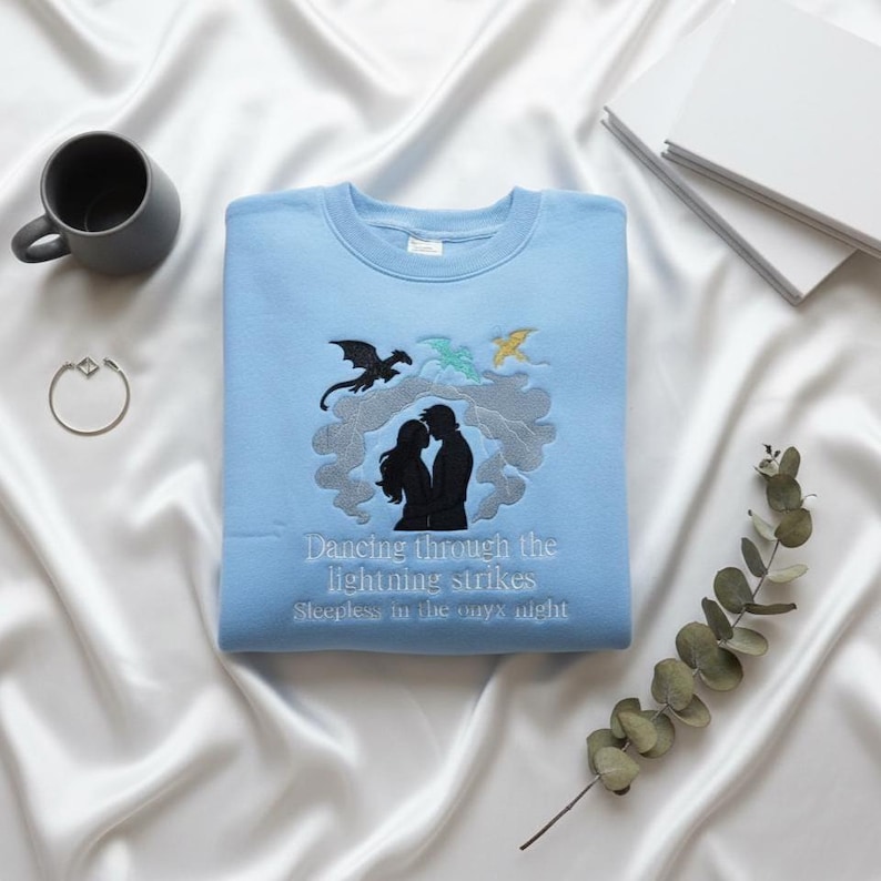 May include: A light blue sweatshirt with embroidered design. The design features a silhouette of a couple, dragons, and the text "Dancing through the lightning strikes, Sleepless in the onyx night."