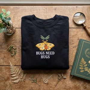 May include: A black crewneck sweatshirt featuring an embroidered design of a yellow moth with a small orange beetle on its back, positioned below a green sprig. The text "BUGS NEED HUGS" is embroidered in white capital letters below the moth.