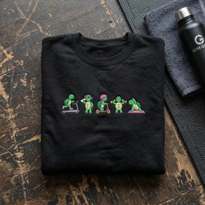 May include: Black crewneck sweatshirt featuring a line of cartoon turtles engaged in various fitness activities. The turtles are depicted on a treadmill, drinking water, riding a scooter, lifting weights, and doing yoga. A black water bottle and a gray towel are in the background.